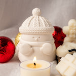 Snowman-shaped candle holder with a lit candle surrounded by Christmas ornaments on a soft surface.