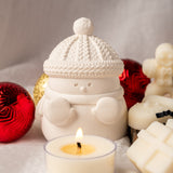 Snowman-shaped candle holder with a lit candle surrounded by Christmas ornaments on a soft surface.