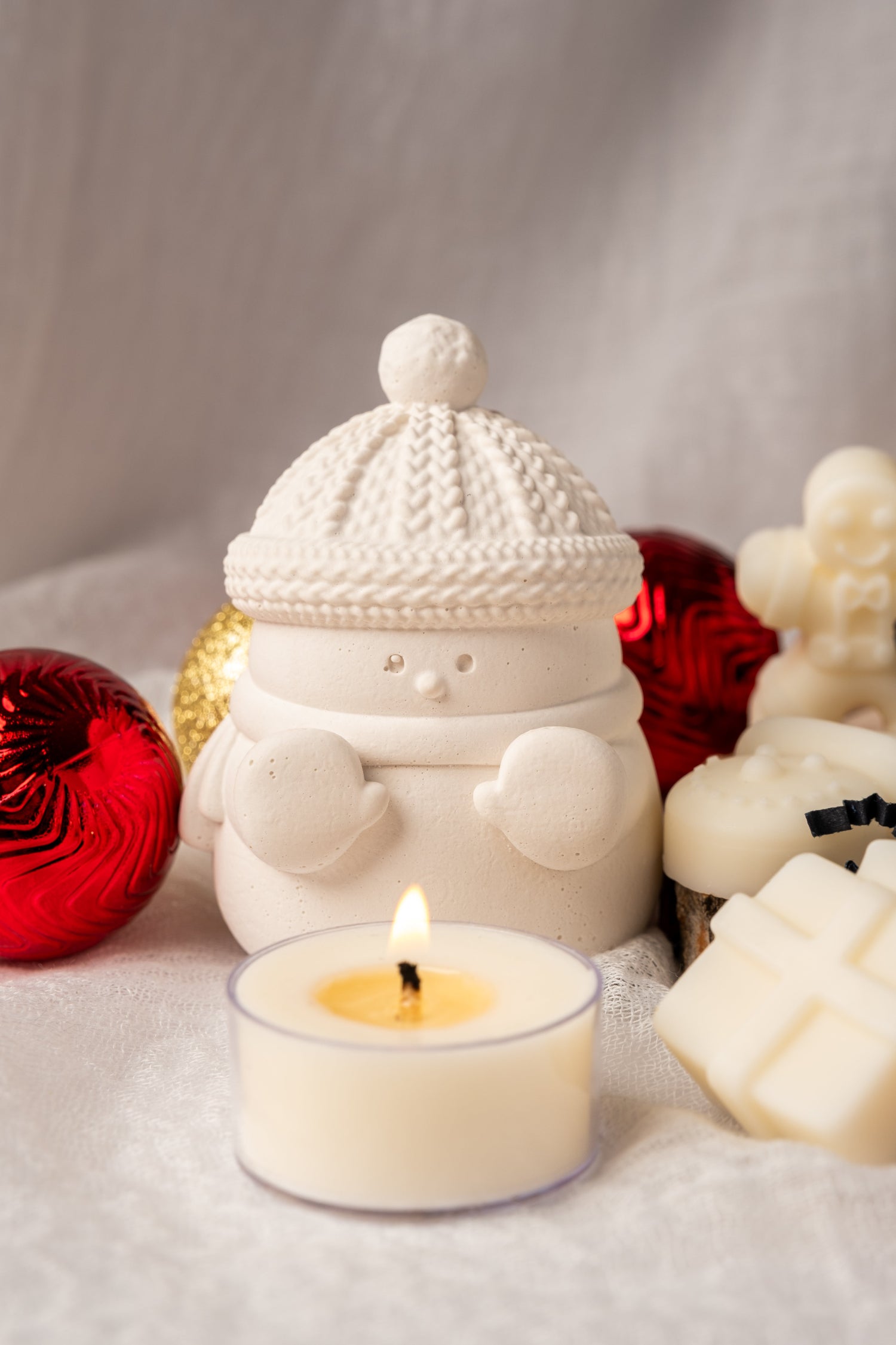 Snowman-shaped candle holder with a lit candle surrounded by Christmas ornaments on a soft surface.