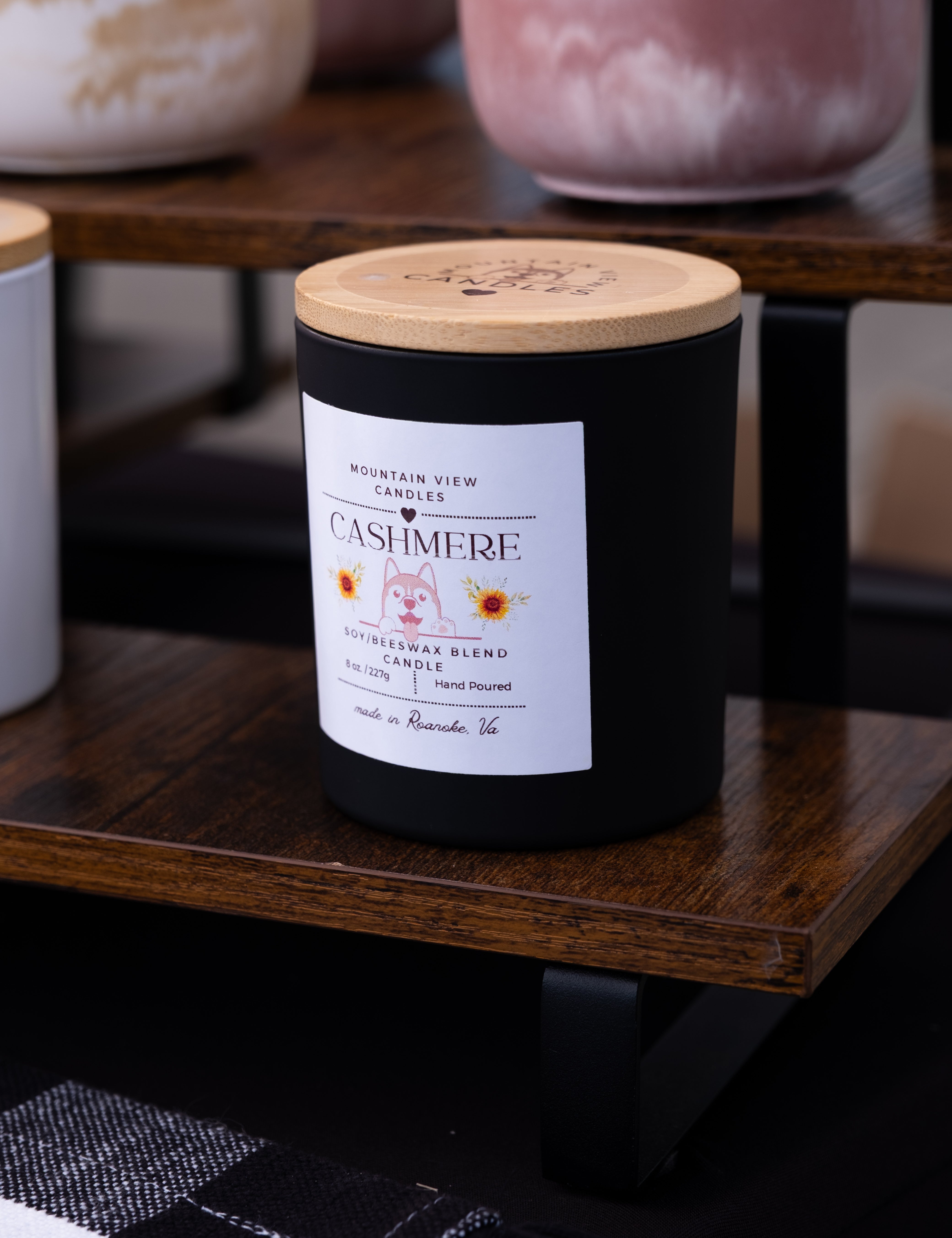 Black candle with a wooden lid on a wooden shelf, featuring a label.