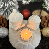 Decorative candle holder with two white figures wearing hats, surrounded by Christmas ornaments.
