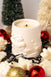 White candle in a decorative holder surrounded by Christmas ornaments on a light background