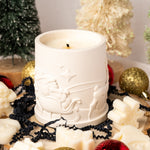 White candle with decorative design surrounded by Christmas ornaments and decorations.